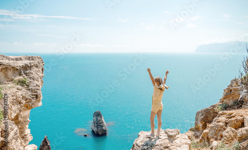 Happy girl stands on a rock high above the sea, wearing a yellow jumpsuit and sporting braided hair, depicting the idea of a summer vacation by the sea.
