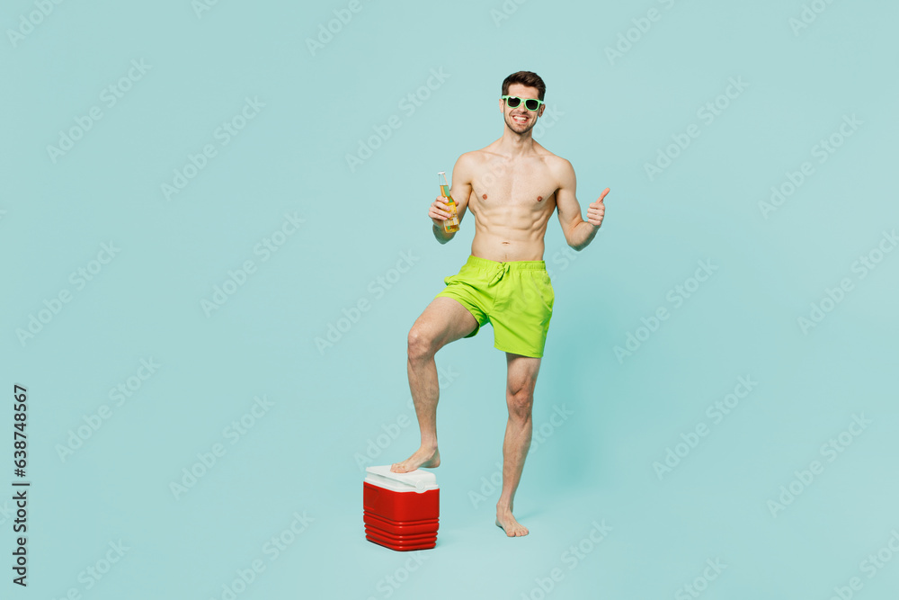 Full body cheerful happy fun young man wear green shorts swimsuit relax near hotel pool with fridge box beer show thumb up isolated on plain blue background. Summer vacation sea rest sun tan concept.