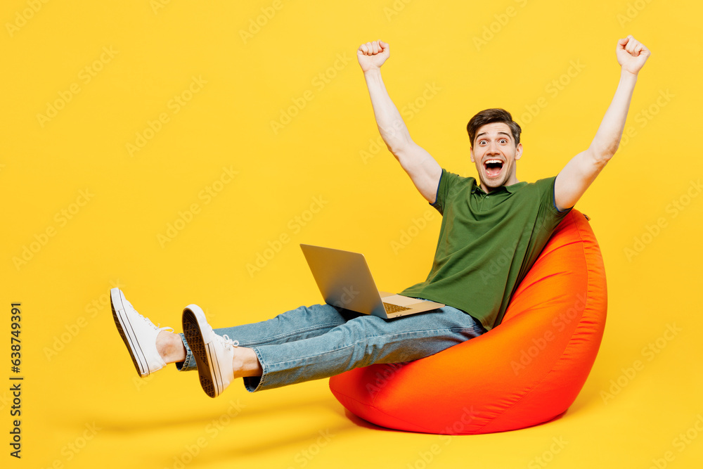 Full body young happy IT man he wears green t-shirt casual clothes sit in bag chair hold use work on laptop pc computer do winner gesture isolated on plain yellow background studio. Lifestyle concept. Full body young happy IT man he wears green t-shirt casual clothes sit in bag chair hold use work on laptop pc computer do winner gesture isolated on plain yellow background studio. Lifestyle concept.