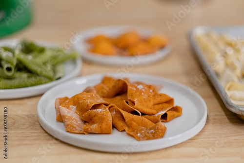 Closeup of handmade pasta, cut into thick, wide, noodles and brightly colored with tomato paste. Other shapes and colors of handmade pasta fill the background.