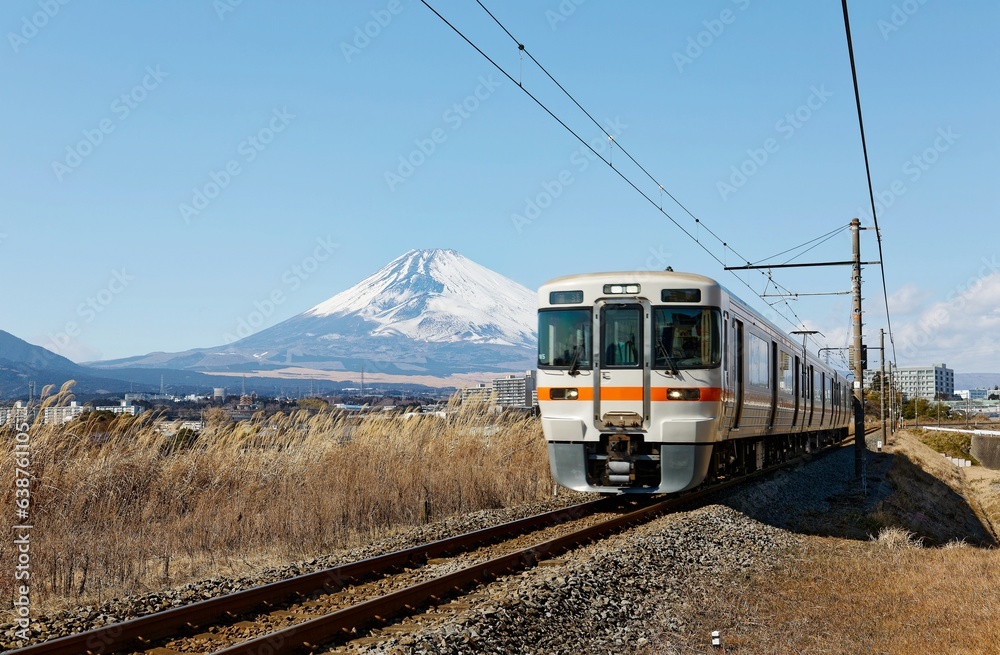 A local train of JR Gotemba Line (御殿場線) traveling through the countryside on a sunny winter day and snow capped Mount Fuji dominating the background under blue clear sky in Susono, Shizuoka, Japan