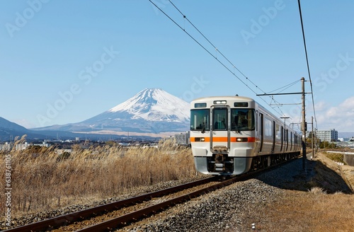 A local train of JR Gotemba Line (御殿場線) traveling through the countryside on a sunny winter day and snow capped Mount Fuji dominating the background under blue clear sky in Susono, Shizuoka, Japan photo