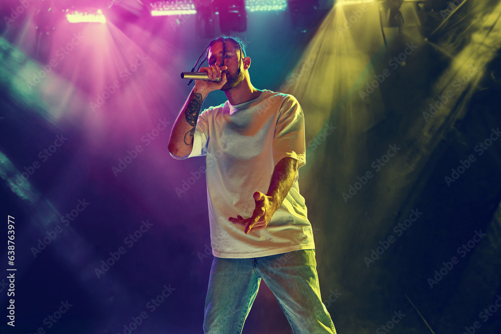 Young artistic man, musician making live concert, singing, dancing on stage over colorful neon illumination. Night club. Music, performance, art, talent, nightlife, joy, party and lifestyle concept