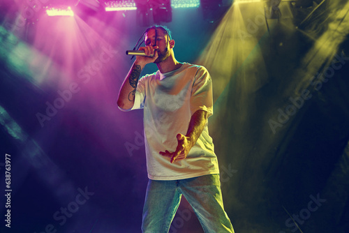 Young artistic man, musician making live concert, singing, dancing on stage over colorful neon illumination. Night club. Music, performance, art, talent, nightlife, joy, party and lifestyle concept