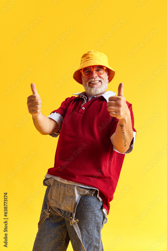 Half-length portrait of happy satisfied senior man showing thumb up. Beard and denim, jeans suit, red t-shirt, glasses and hat. Isolated on yellow. Half-length portrait of happy satisfied senior man showing thumb up. Beard and denim, jeans suit, red t-shirt, glasses and hat. Isolated on yellow.