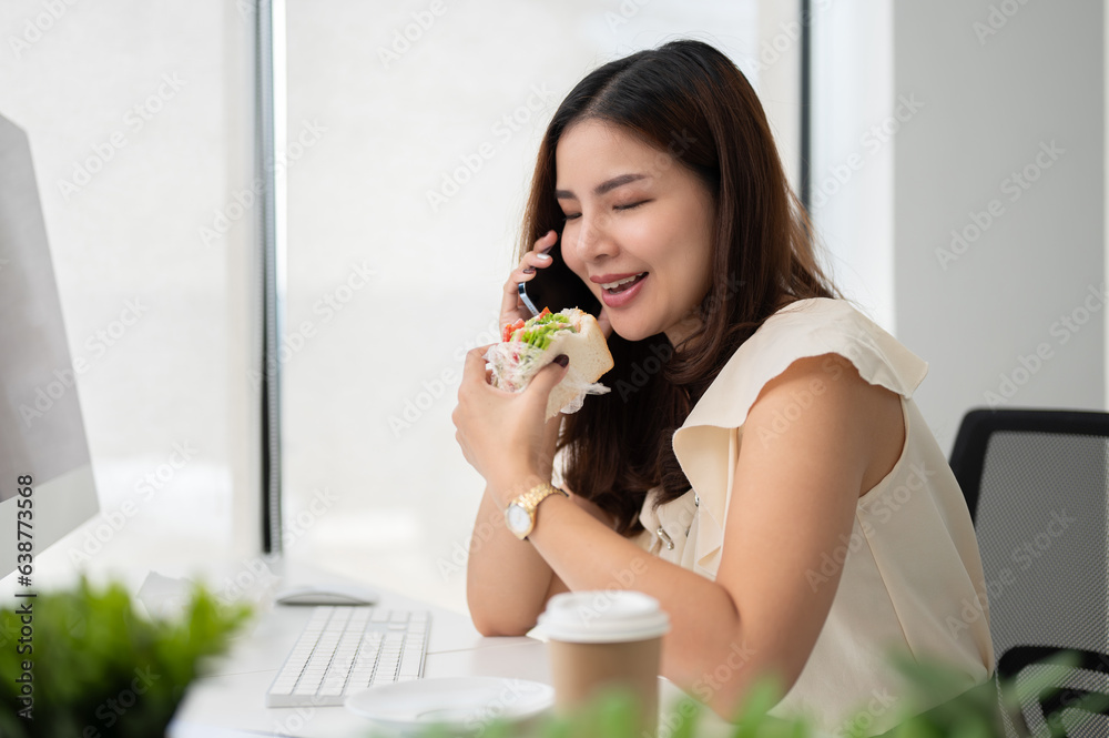 A happy Asian businesswoman is eating a sandwich and talking on the phone at her desk A happy Asian businesswoman is eating a sandwich and talking on the phone at her desk