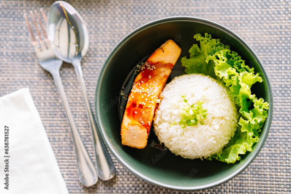 Steamed rice with grilled salmon in a black bowl and spoon on the dining table in a restaurant.