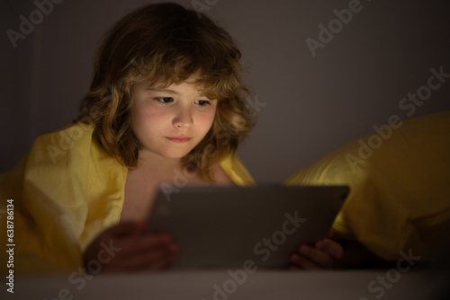 Kid playing on a digital tablet. Kid using tablet for gaming and online learning while lying on the bed, screen light reflex on her face. Child play tablet under blanket at night in a dark room. © Volodymyr