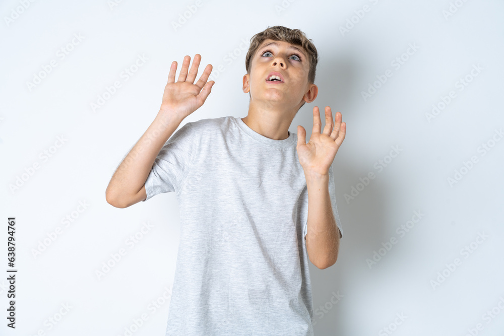 Beautiful kid boy wearing grey casual t-shirt keeps palms forward and looks with fright above on ceiling tries to defense herself from invisible danger opens mouth. Beautiful kid boy wearing grey casual t-shirt keeps palms forward and looks with fright above on ceiling tries to defense herself from invisible danger opens mouth.