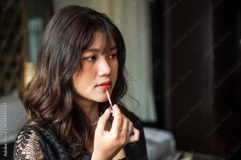 Beautiful young smiling woman applying bright red lipstick cream while looking into the mirror in hotel room. Confident asian girl looking at her reflection while using liquid lipstick to her mouth.