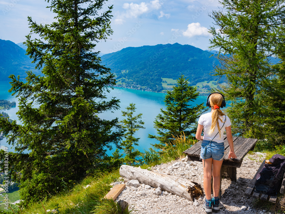 Naklejka premium A teenage girl in headphones stands near a bench on a mountain with Lake Attersee in the background, Austria.