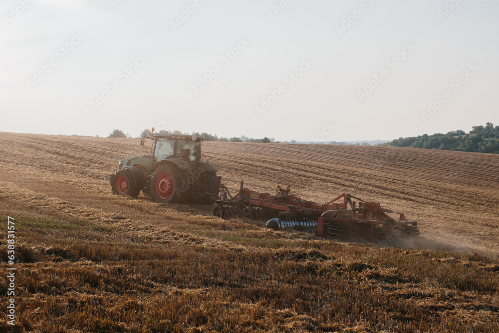 Obraz premium A modern tractor with a heavy trailed disc harrow plows a wheat field at sunset.