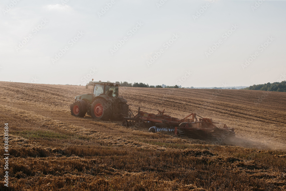 Obraz premium A modern tractor with a heavy trailed disc harrow plows a wheat field at sunset.