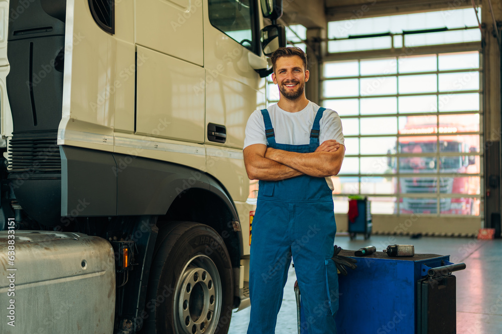 Portrait of professional truck mechanic standing in vehicle workshop. In background parked trucks ready to be serviced.