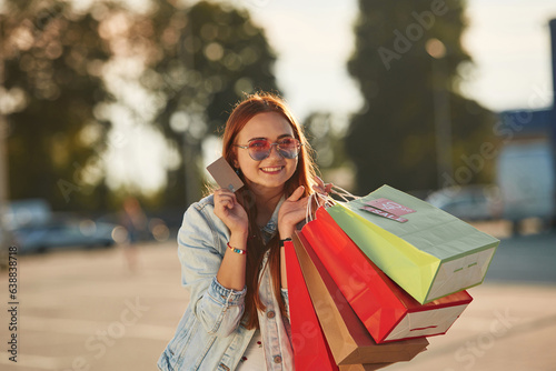 Discounts, with credit card in hands. Beautiful woman in casual clothes is holding shopping bags, outdoors