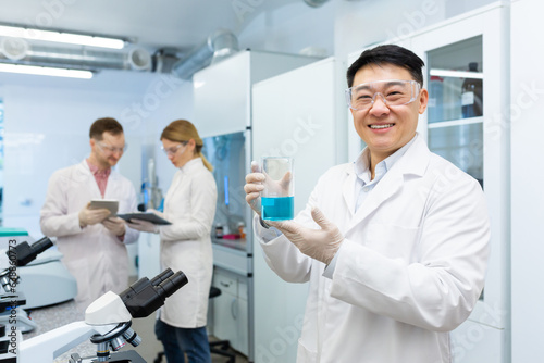 Portrait of a young smiling Asian man, a medical student standing in a laboratory and holding a flask with blue liquid in his hands, behind colleagues discussing achievements and results.