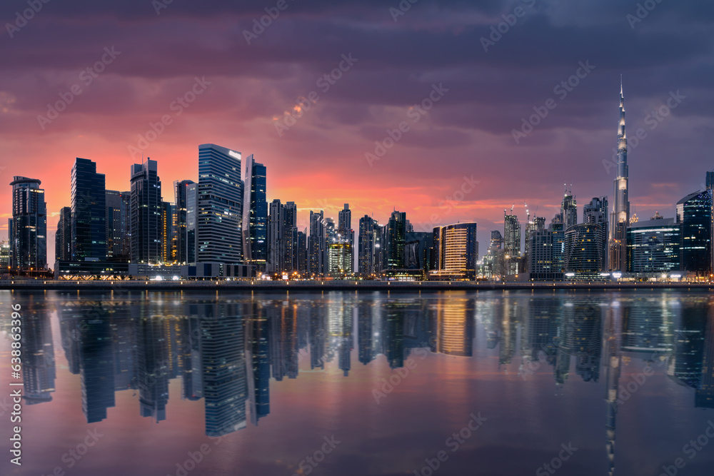 Stunning view of the illuminated Dubai skyline at sunset with the magnificent Burj Khalifa and buildings reflected on a silky smooth water in the foreground.