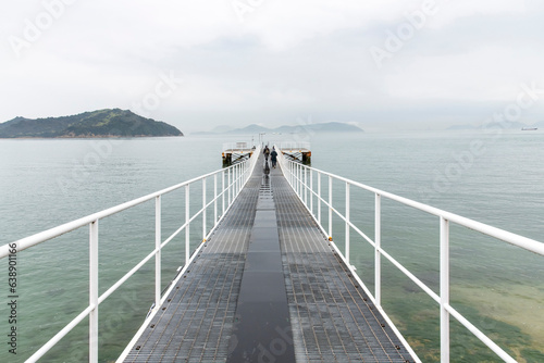 View of two people walking the length of a jetty or pier into the Seto Inland Sea of Japan near Naoshima Island with foggy and misty conditions and overcast partly covering mountains