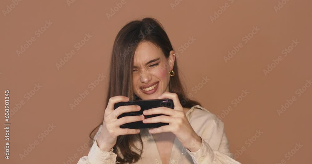 Happy modern brunette girl using mobile phone, young woman playing video game on smartphone, standing isolated over beige background. Happy modern brunette girl using mobile phone, young woman playing video game on smartphone, standing isolated over beige background.