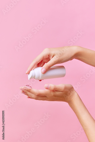 Vertical cropped photo of female hand squeezing cream, gel for hands from unmarked jar with dispenser on palm. Close-up, pink background. © Lustre