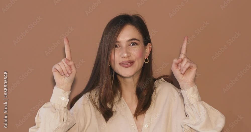 Attractive brunette woman looking at camera and pointing index finger up at copy space for for advertising with smile and positive emotions, excited, look up isolated on beige background. Attractive brunette woman looking at camera and pointing index finger up at copy space for for advertising with smile and positive emotions, excited, look up isolated on beige background.