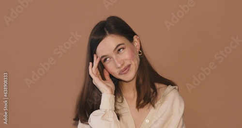 Shocked shy charming confused young woman in beige shirt posing isolated on beige background studio. Girl looking at camera smile hold hands on head, cover face.