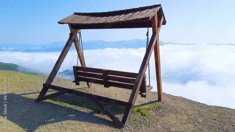A swing swings on a mountain top over an abyss in front of beautiful clouds.