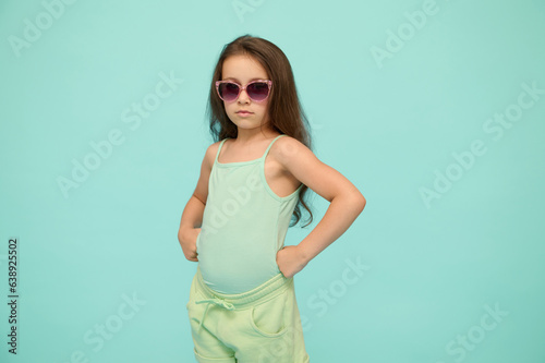 Emotional authentic confident strong kid girl in sunglasses, puts her hands on waist and looking confidently at camera