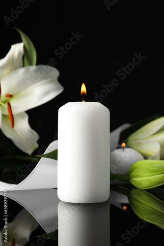 White lilies and burning candle on black mirror surface in darkness  space for text. Funeral symbols