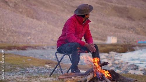 Indian male tourist sitting besides the bonfire in front of the mountain in Zanskar Valley at Ladakh, India. Tourist warming himself with fire in cold Himalayas in India. Camping background. photo