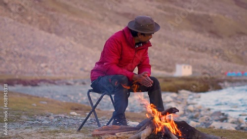 Indian male tourist sitting besides the bonfire in front of the mountain in Zanskar Valley at Ladakh, India. Tourist warming himself with fire in cold Himalayas in India. Camping background. photo