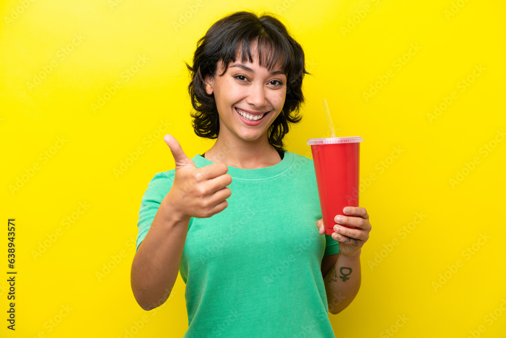 Young Argentinian woman holding a soda isolated on yellow background with thumbs up because something good has happened Young Argentinian woman holding a soda isolated on yellow background with thumbs up because something good has happened