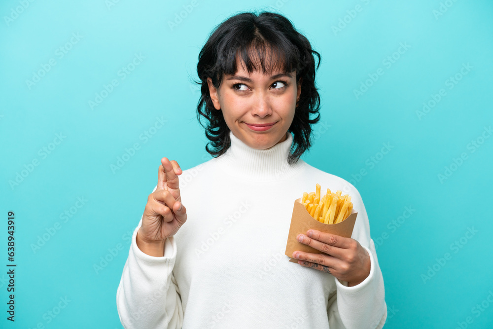 Young Argentinian woman holding fried chips isolated on blue background with fingers crossing and wishing the best Young Argentinian woman holding fried chips isolated on blue background with fingers crossing and wishing the best