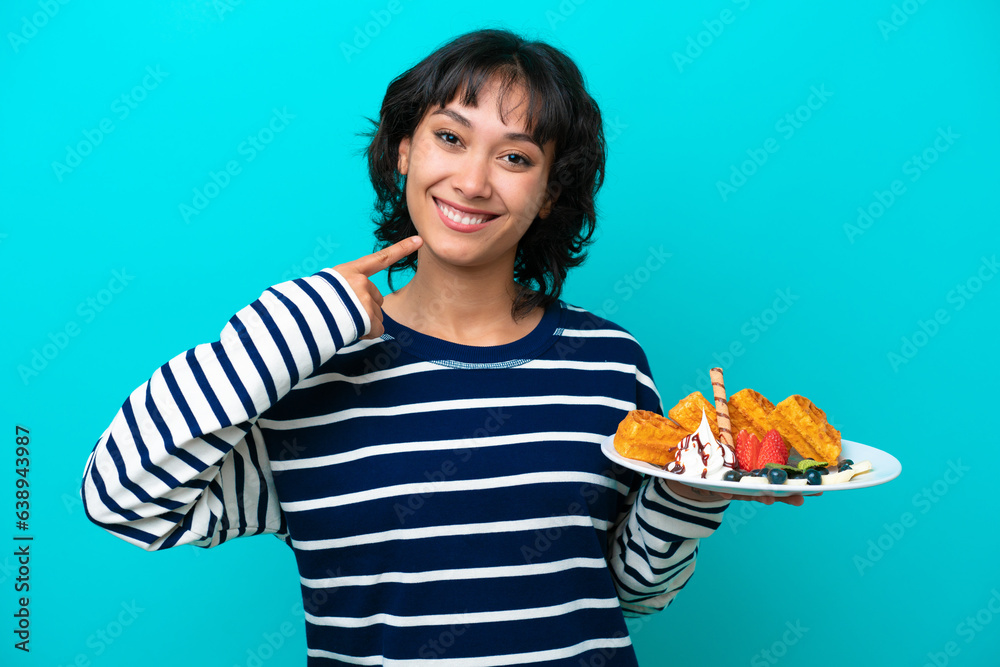 Young Argentinian woman holding waffles isolated on blue background giving a thumbs up gesture Young Argentinian woman holding waffles isolated on blue background giving a thumbs up gesture