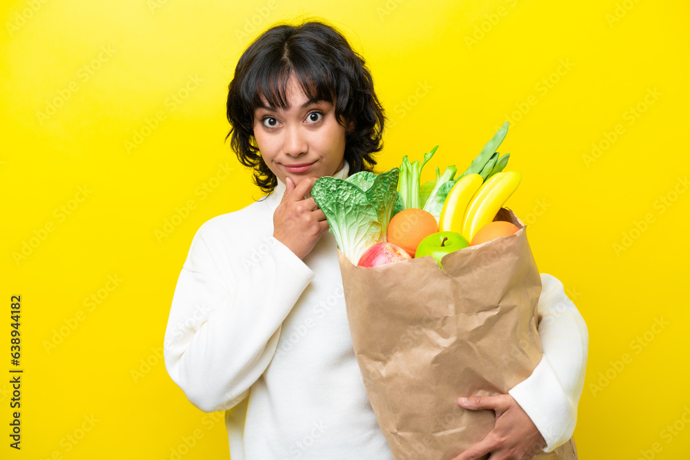 Young Argentinian woman holding a grocery shopping bag isolated on yellow background thinking Young Argentinian woman holding a grocery shopping bag isolated on yellow background thinking