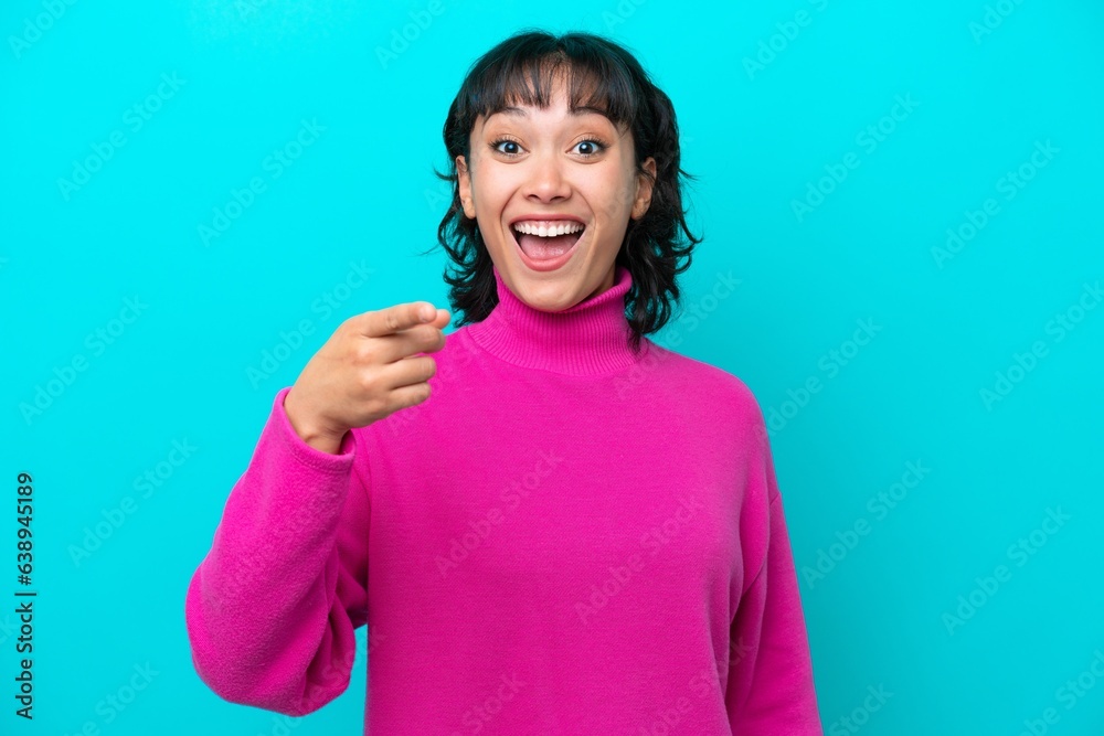 Young Argentinian woman isolated on blue background surprised and pointing front Young Argentinian woman isolated on blue background surprised and pointing front