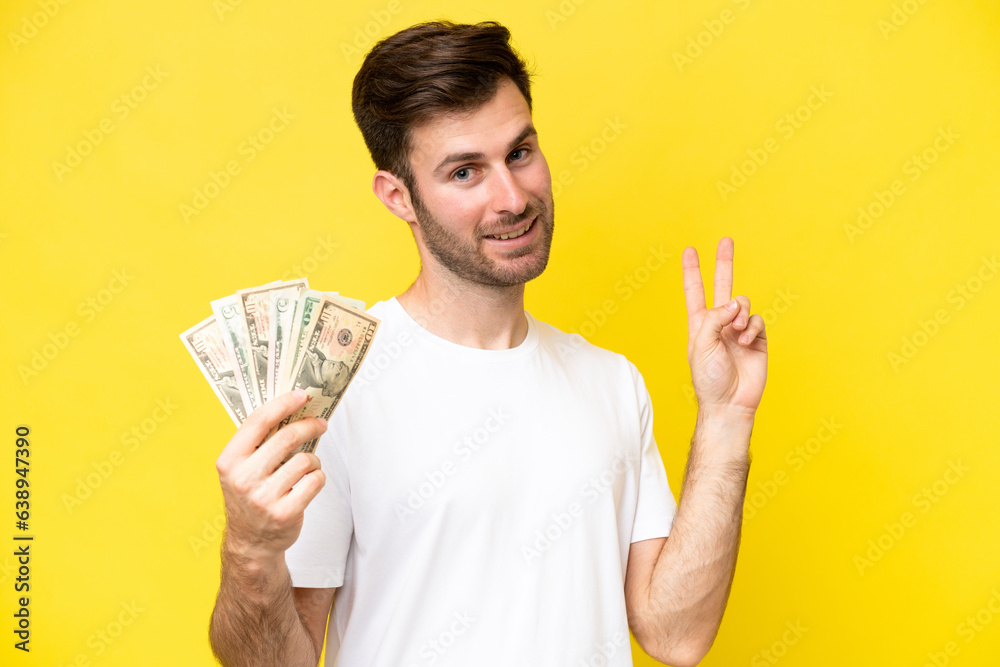 Young caucasian man taking a lot of money isolated on yellow background smiling and showing victory sign Young caucasian man taking a lot of money isolated on yellow background smiling and showing victory sign