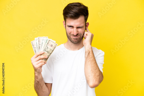 Young caucasian man taking a lot of money isolated on yellow background frustrated and covering ears