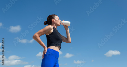 Photos sportswoman drinking water from a glass bottle after exercising outside, blue sk