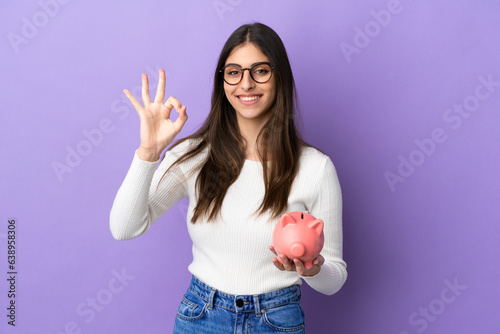 Young caucasian woman holding a piggybank isolated on purple background showing ok sign with fingers
