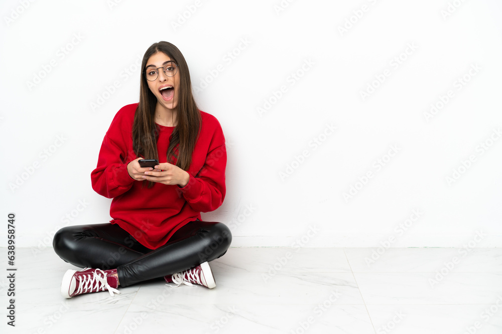 Young woman sitting on the floor isolated on white background surprised and sending a message Young woman sitting on the floor isolated on white background surprised and sending a message