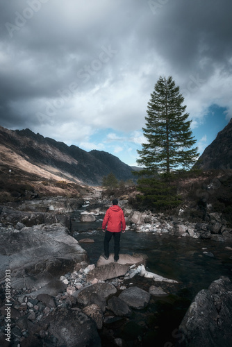 A hiker in a red jacket on the bank of a mountain stream looking out over a magnificent mountain landscape. Scottish Highlands