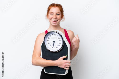 Young reddish woman isolated on white background with weighing machine and doing victory gesture