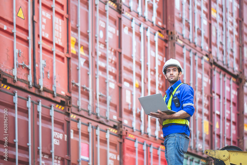 Engineer or cargo worker wears PPE standing in front of cargo stacked container yard and holding laptop and looking at the sky. concept logistic delivery management for industrial.