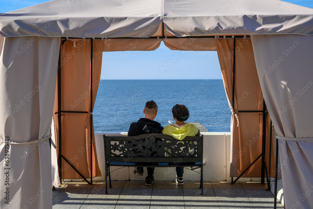 A young guy and a girl are sitting on a bench under an awning and looking at the sea
