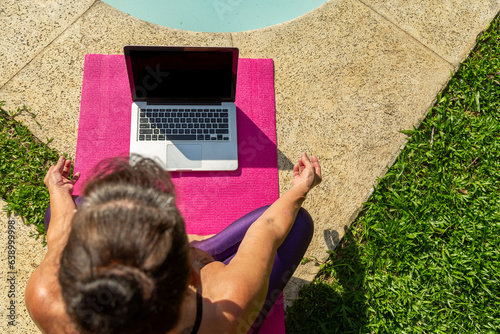 Mulher vista de cima, sentada sobre um tapete rosa de yoga na beira da piscina e gramado ao lado e com seu notebook em sua frente, em dia de sol. photo