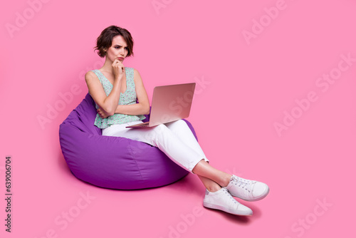 Full body photo of smart woman pouted lips thoughtful hmm watching stats finance results laptop sit pouf isolated on pink color background