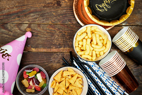 birthday or christmas, new year's party concept, paper plate with card celebrate text and disposable cup, straws and colorful with different chips and candies, pink hat tinsel on wooden table