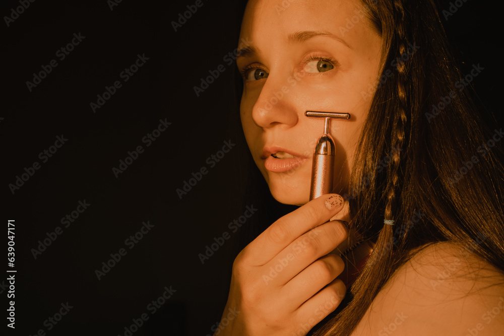 Obraz premium Young beautiful white woman, girl is using vibrating golden facial massager on dark black background with copy space. Preservation prolongation of natural beauty youth Female skin care with pleasure.