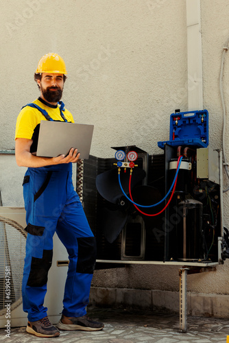 Portrait of smiling knowleadgeable engineer working on external hvac system, holding laptop. Professional wireman optimizing condenser performance, ensuring it operates at maximum capacity photo